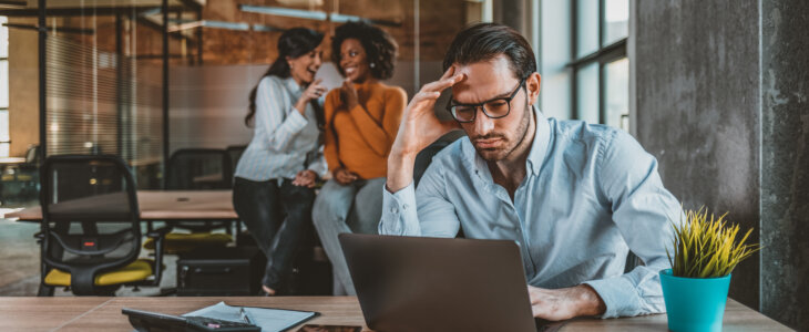 stressed man at laptop while coworkers gossip behind him