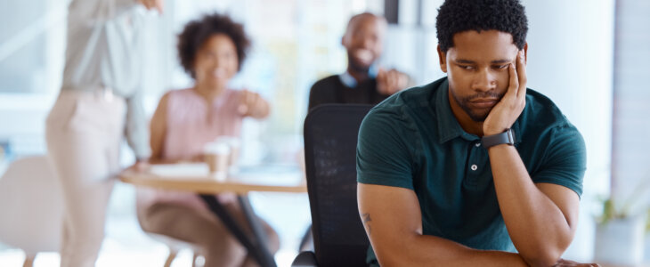 man sitting at desk looking down while coworkers point and laugh in background